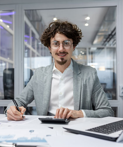 Hombre con gafas y blazer gris escribiendo junto a una calculadora, con un portátil sobre la mesa